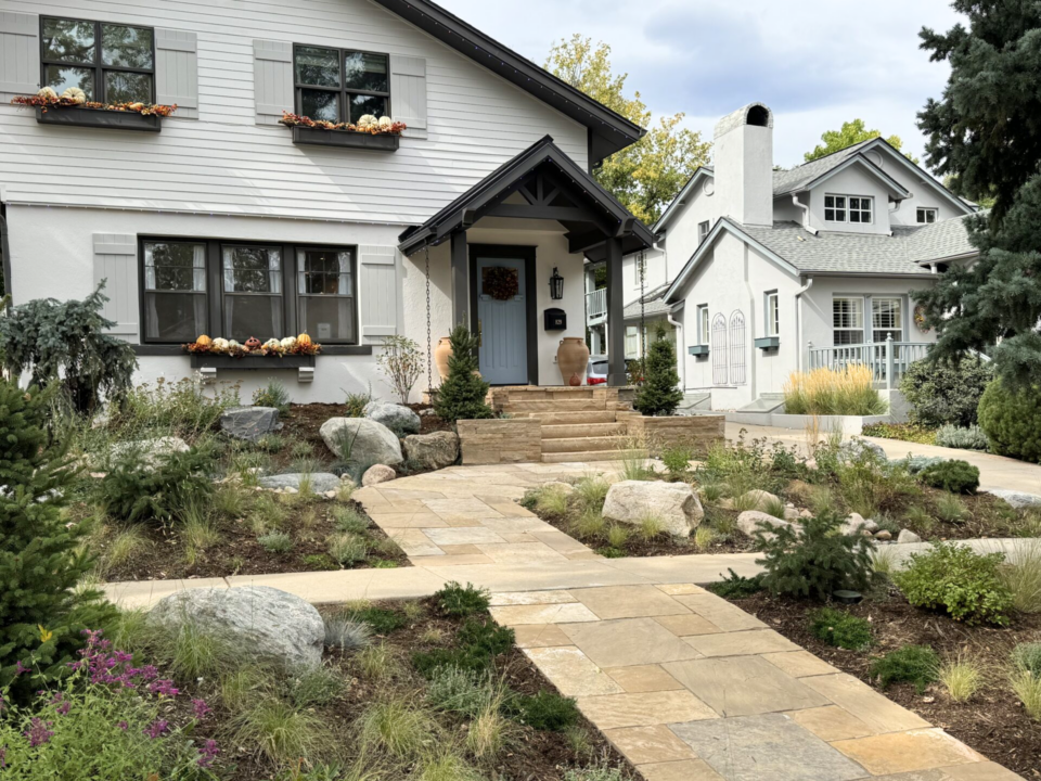 A modern white two-story house with pumpkins on the window sills, a stone walkway, landscaped front yard with rocks, small shrubs, and another similar house in the background.