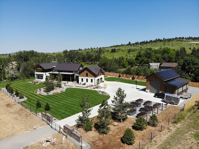 Aerial view of a modern house with a large green lawn, landscaped garden, driveway, and a separate garage or outbuilding with solar panels, set against a backdrop of rolling hills and trees under a clear blue sky.