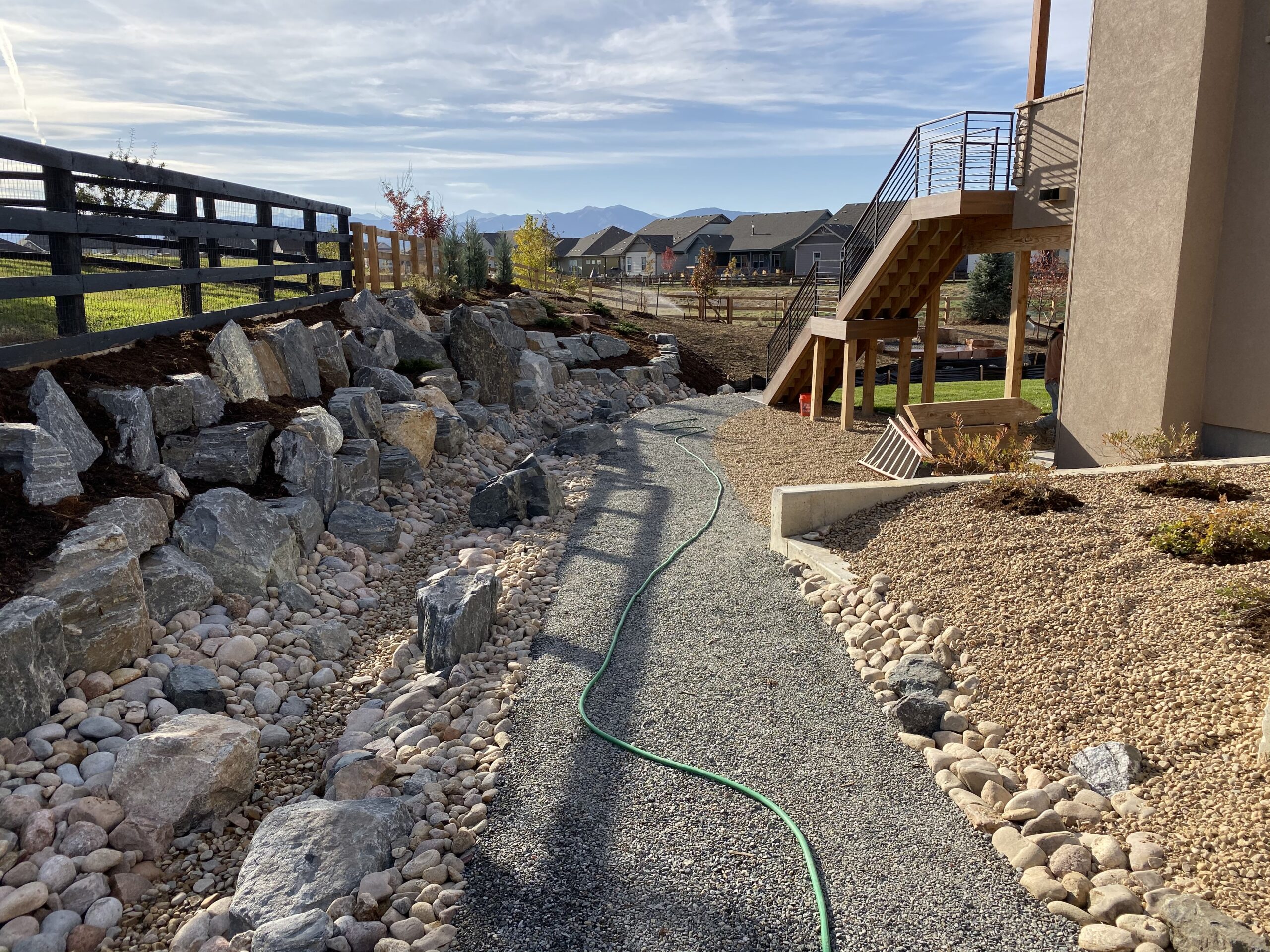 A gravel pathway bordered by rocks and dry landscaping curves beside a house with an outdoor staircase. In the background are fenced yards, houses, and mountains under a blue sky. A green garden hose lies on the path.