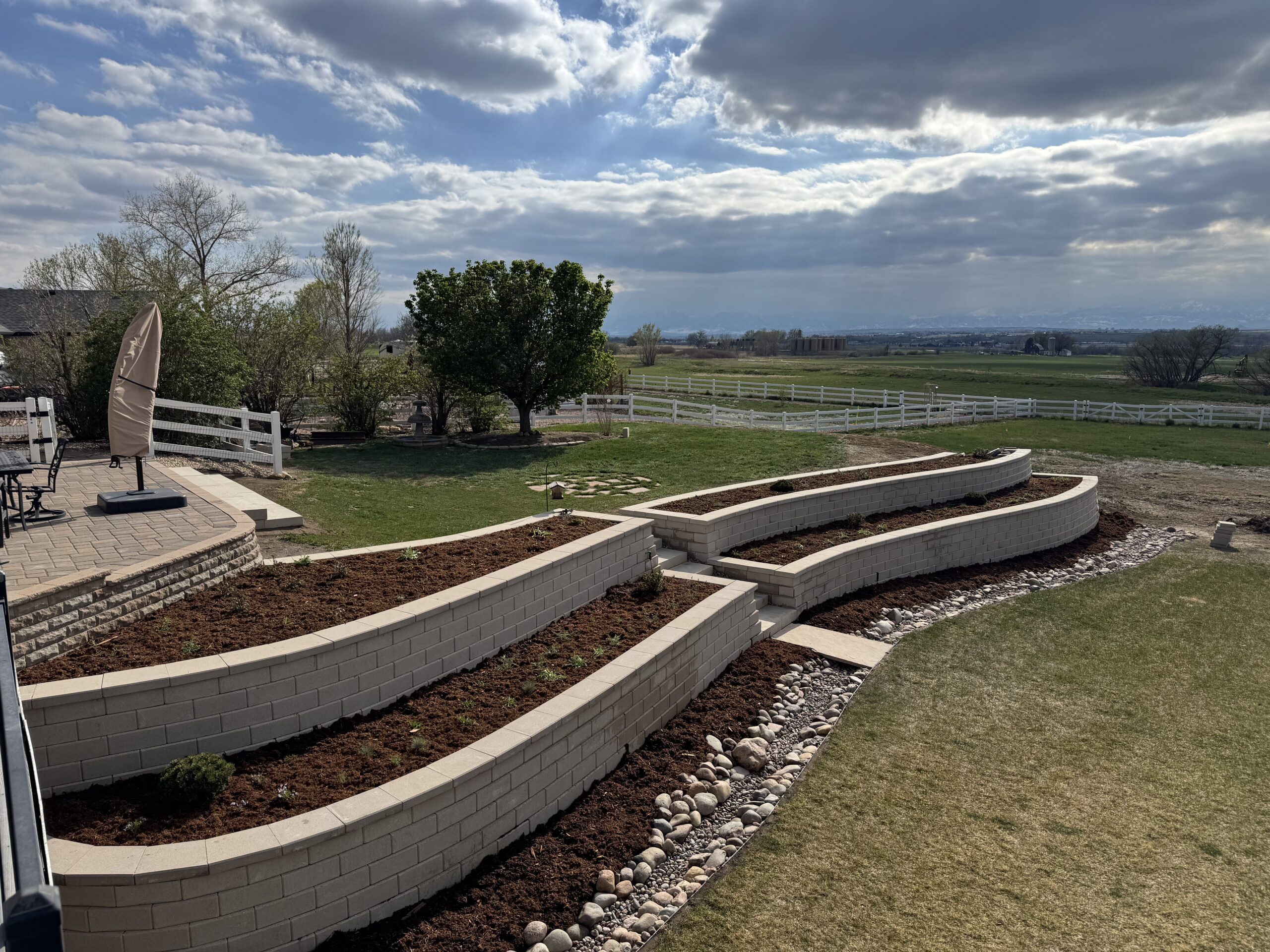 A backyard with tiered stone garden beds filled with soil and a border of rocks, a patio with an umbrella, green lawn, trees, and distant open fields under a partly cloudy sky.
