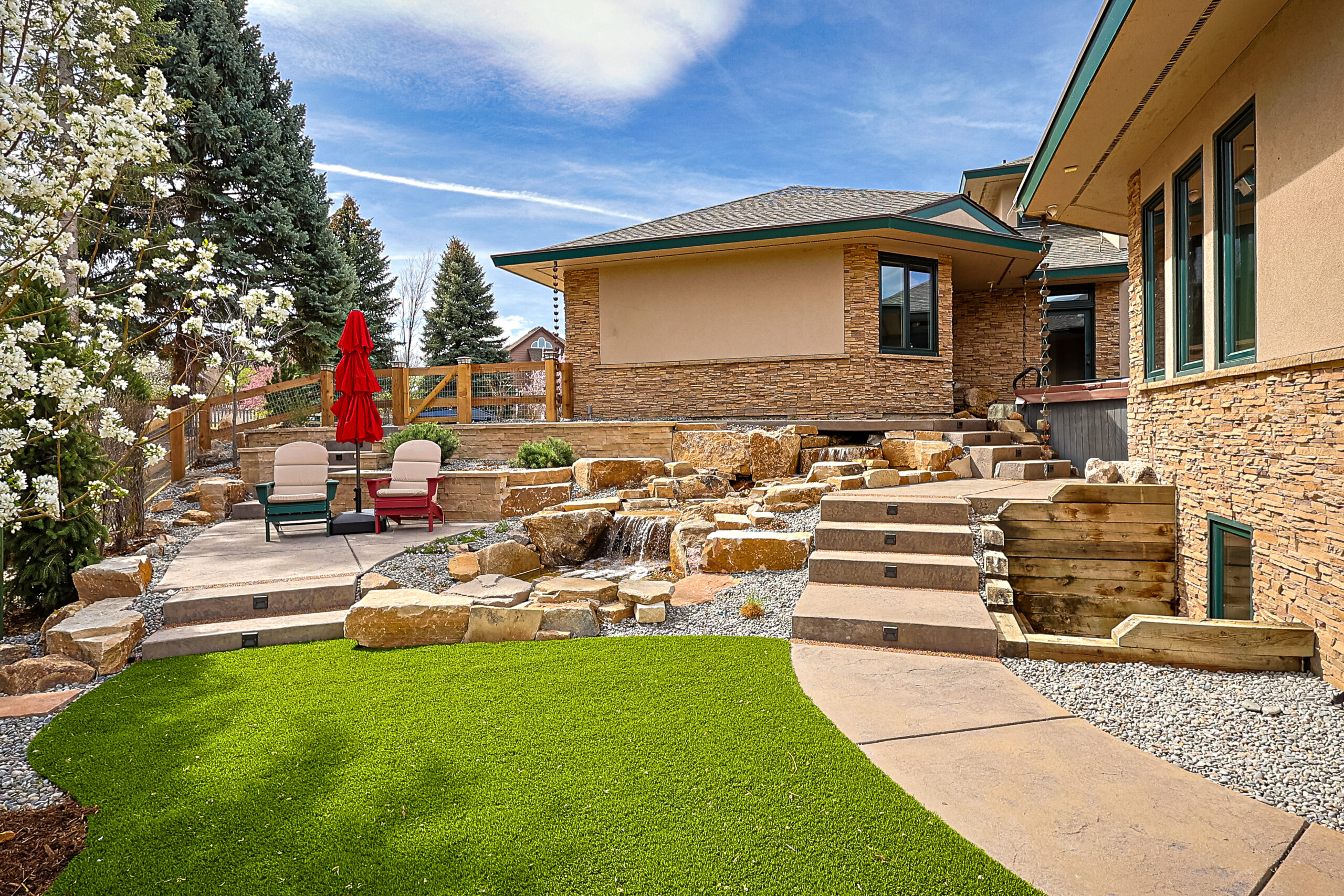 A landscaped backyard with artificial grass, stone steps, a waterfall feature, two lounge chairs, a red umbrella, and a tan house with stone accents under a partly cloudy sky.